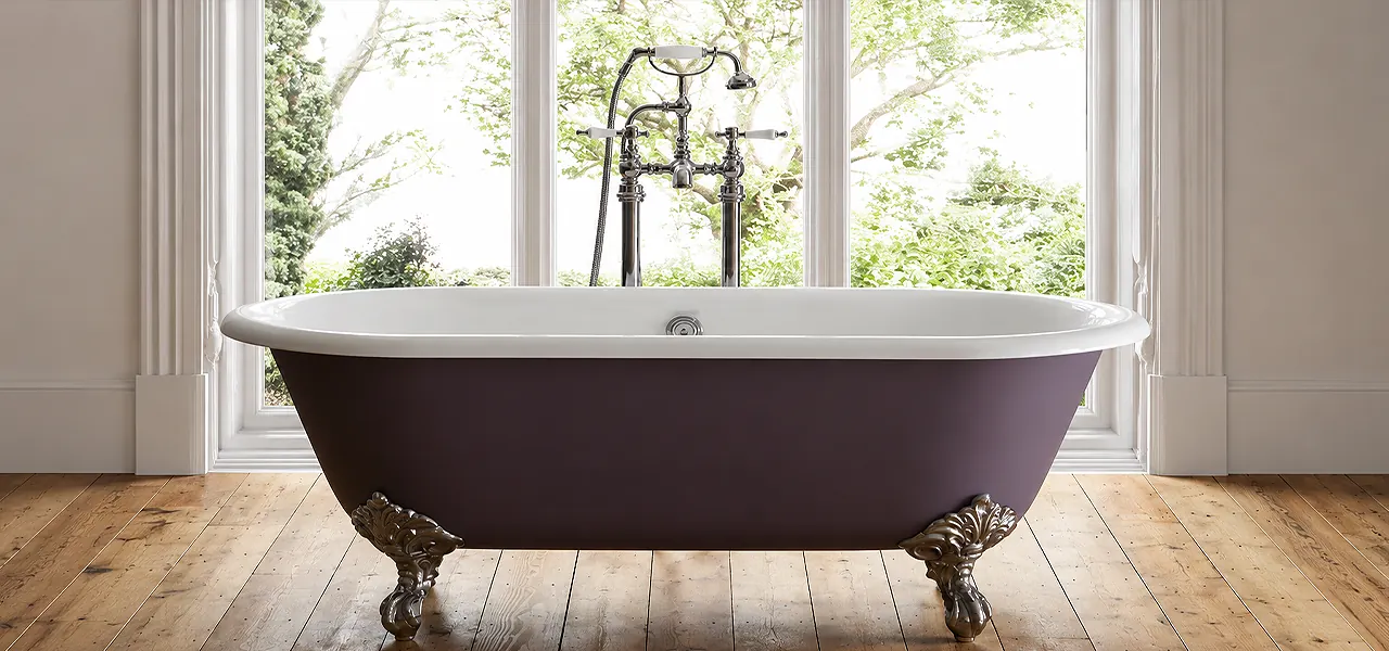 Aubergine cast iron roll-top freestanding bath with polished metal claw feet and chrome telephone taps, in front of a large garden window on a wood floor.
