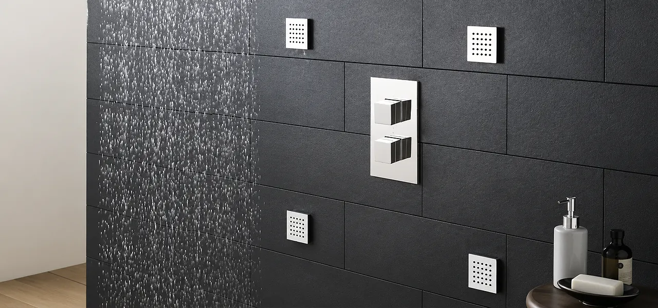 Several square chrome body jets and a thermostatic shower valve on a dark tiled wall, with a rainfall shower spray running alongside.