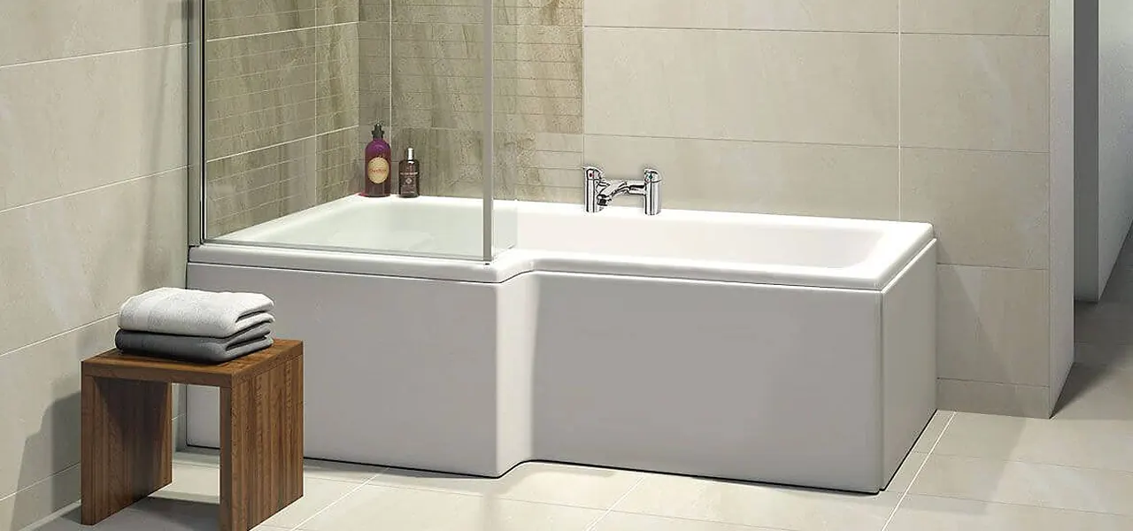 White L-shaped shower bath with a single fixed glass screen panel and deck-mounted chrome taps, in a beige-tiled bathroom with a wooden stool.