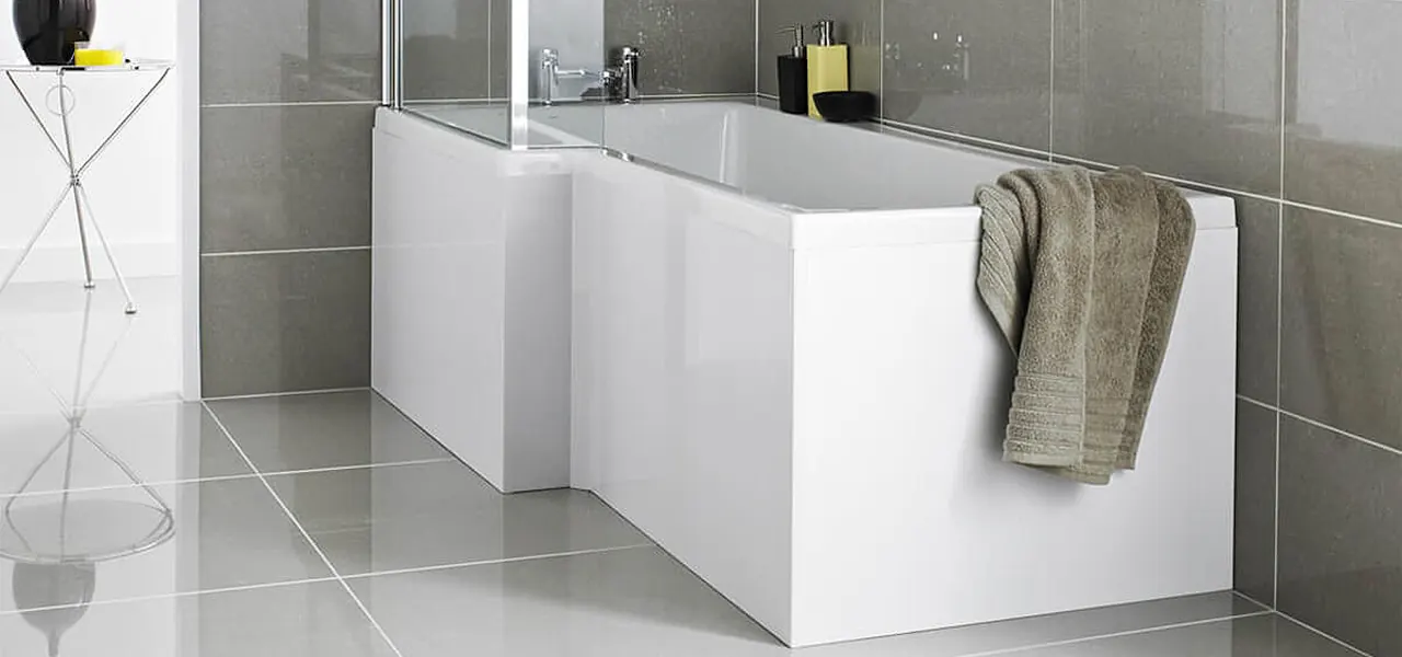 White L-shaped shower bath fitted with gloss front and end panels, a taupe towel draped over the rim, in a grey-tiled bathroom.