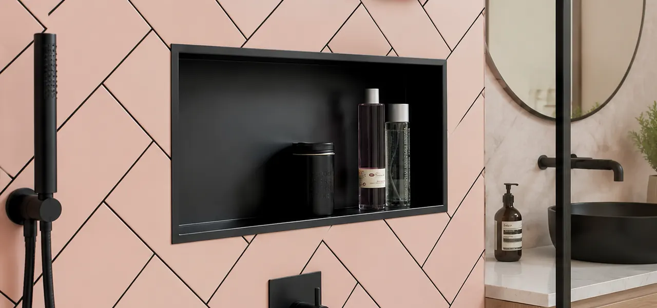 A rectangular matt black recessed niche set into a wall of blush pink herringbone tiles with black grout lines, holding three glass toiletry bottles and a small canister, with matt black shower