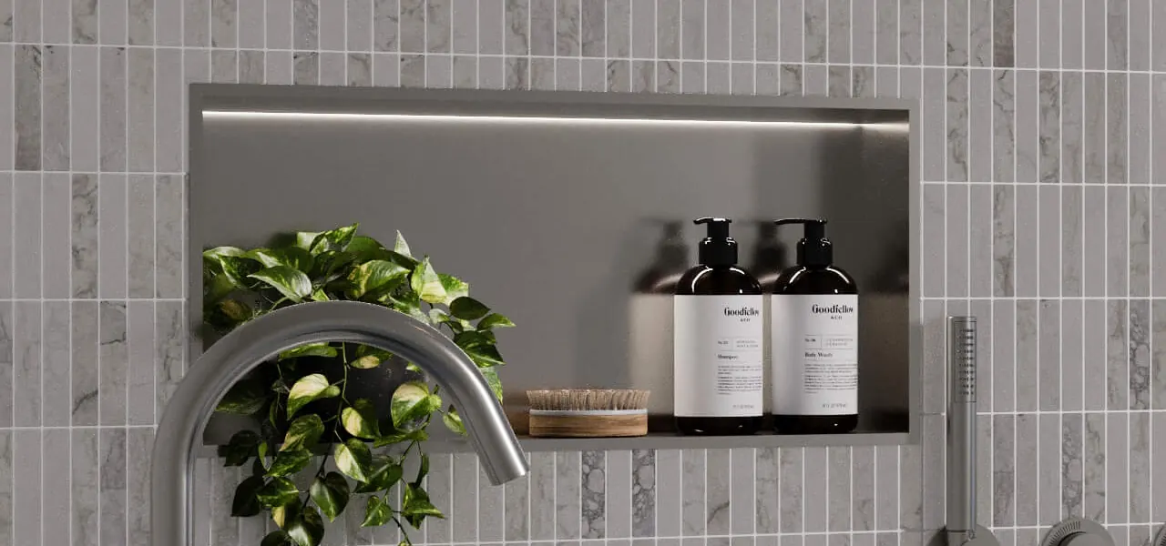 A horizontal stainless steel shower niche set into a wall of gray vertical mosaic tiles, holding two amber pump bottles of shampoo and body wash and a wooden nail brush, with a trailing pothos plant draped beside a chrome tap.