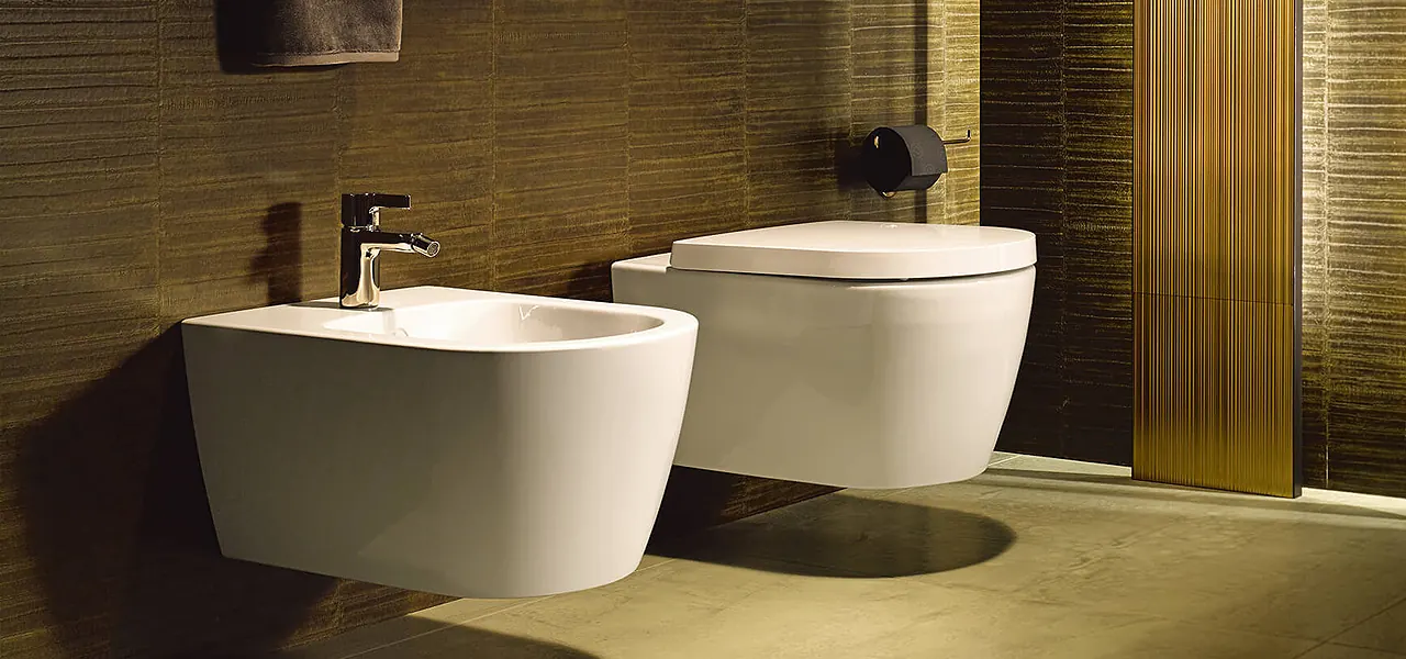 White wall-hung toilet and matching bidet with chrome and matt black fittings, against warm earthy textured tiles.