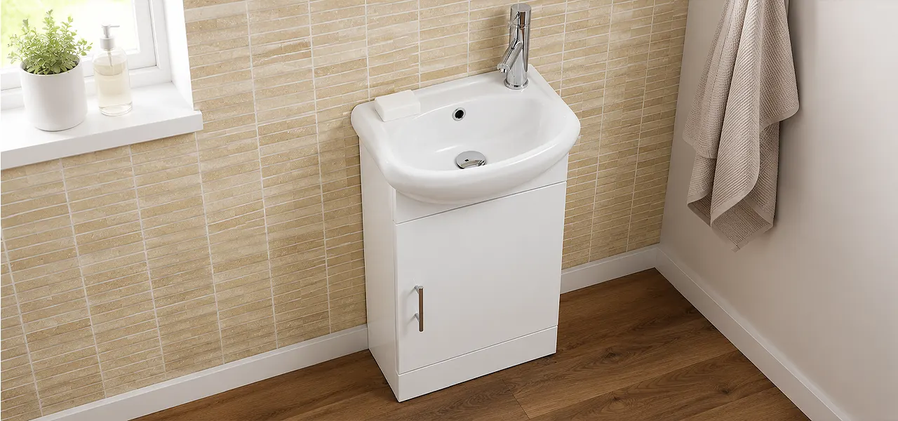 White floor-standing cloakroom vanity unit with a rounded compact basin and chrome tap, against beige brick-effect tiles on a wood floor.