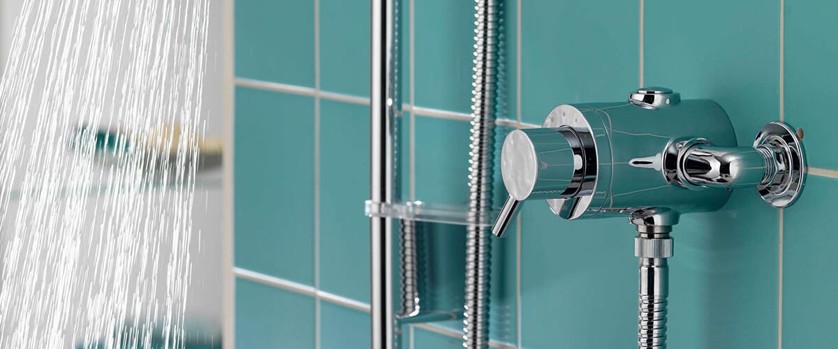 Close-up of a modern shower control with chrome fixtures mounted on teal wall tiles, with water spraying from the showerhead.
