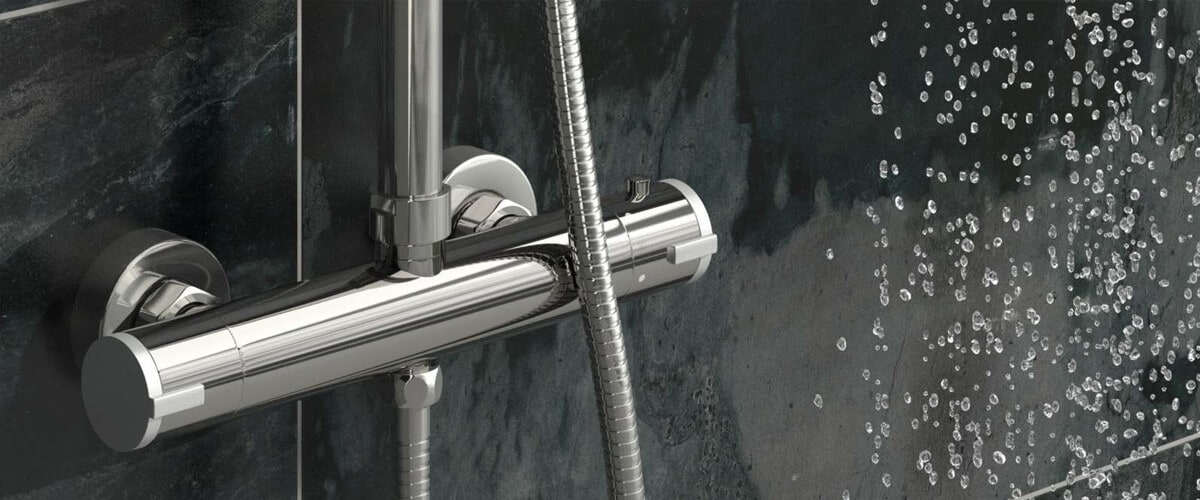 Close-up of a chrome thermostatic shower valve and hose against a dark tiled wall, with water droplets spraying nearby.