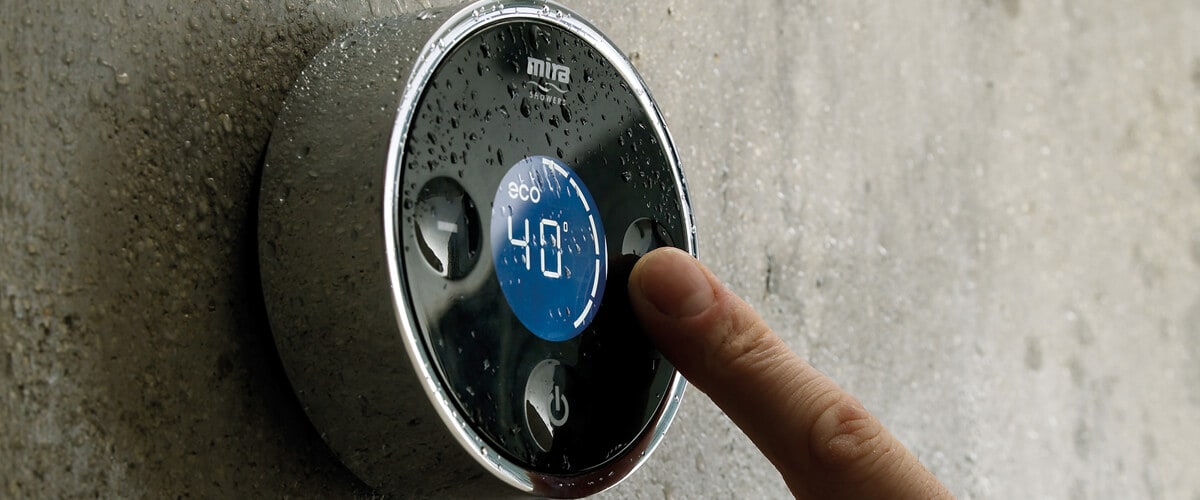 Close-up of a digital shower thermostat with illuminated temperature display, mounted on a wet wall as a hand reaches to adjust the control.