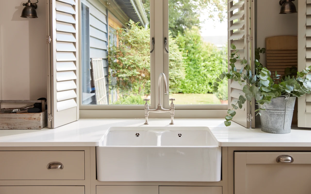 Traditional Belfast farmhouse sink installed beneath a window in a bright kitchen, showcasing a deep apron-front fireclay design with classic cabinetry.